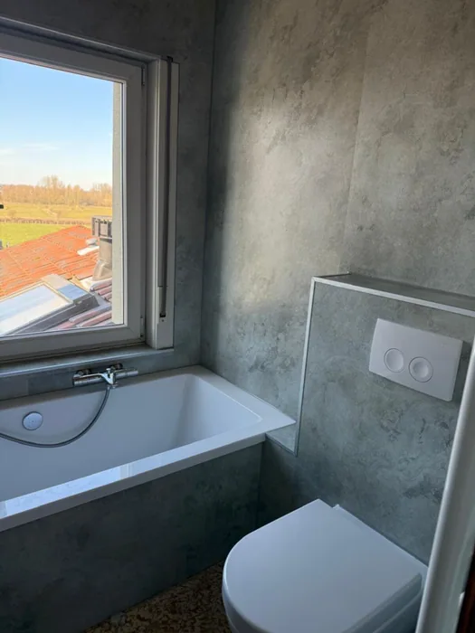 Ligbad onder het raam met grijze betonlook wandpanelen en hangtoilet